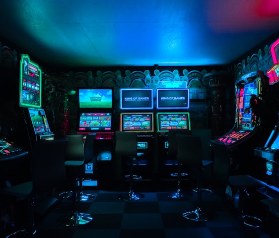 Dimly lit gaming arcade featuring a variety of brightly illuminated slot machines and video gaming screens. Neon signs and colorful displays create a vibrant atmosphere. The arrangement of gaming machines suggests an inviting space for entertainment, with chairs positioned for players.