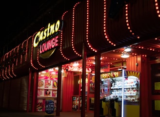 A brightly lit casino lounge entrance adorned with neon lights. The exterior features colorful signage including the words 'Casino' and 'Lounge' in vivid colors, with additional lights framing the building. Inside, a glimpse of a claw machine filled with plush toys and arcade games is visible. The area exudes an inviting yet bustling atmosphere typical of entertainment venues.