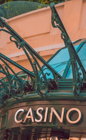 The image displays the entrance to a casino, characterized by detailed and ornate architectural elements with green metal structures and a sign that reads 'CASINO'. The background includes a glass dome and a peach-colored wall.