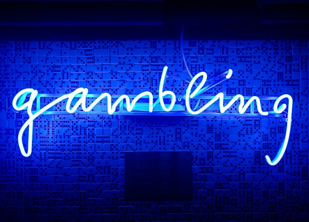 A bright blue neon sign spells out the word 'gambling' in cursive script. The background features a textured wall covered in domino tiles, also illuminated by the blue light, creating a vivid and visually striking scene.