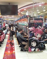 A bustling arcade filled with people playing various gaming machines. Brightly colored displays and animated characters are prominent on posters and screens. The atmosphere is lively with patrons seated in rows, focusing on the games. Overhead signage and electronic scoreboards are visible, contributing to the busy and vibrant environment.