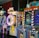 A row of brightly colored arcade game machines is arranged in a line against the wall. Each machine features vibrant lights and designs, with themes ranging from claw games to game show-inspired setups. The area has a playful and inviting atmosphere, suitable for entertainment or amusement centers.