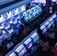 A bustling arcade filled with rows of gaming machines, each with vibrant displays and controls. Several people are seated at these machines, deeply engaged in playing games. Bright fluorescent lights cast a blue and neon glow across the room, enhancing the lively and energetic atmosphere.