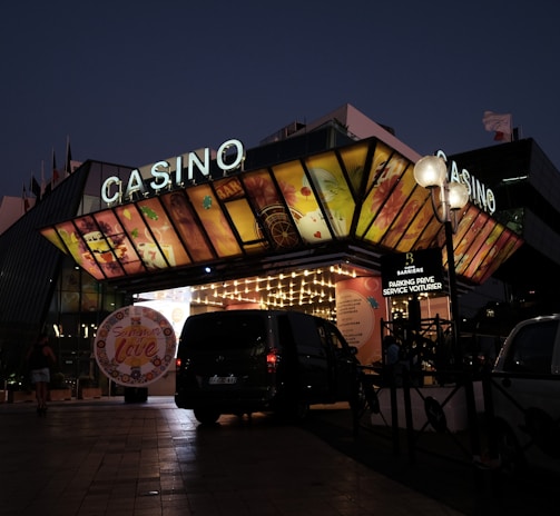 A building with a brightly illuminated entrance is seen at night. The sign above reads 'Casino' in bright letters. The facade features colorful graphics depicting various indulgent themes. Several vehicles are parked near the entrance, and people are visible in the vicinity. Tall lamp posts with spherical lights are present, casting a warm glow.
